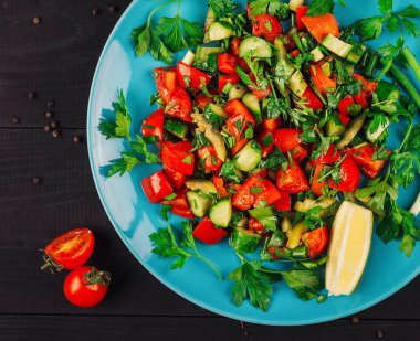 Bitkilerle taze sebze salatası ve siyah ahşap arka planda mavi bir tabakta limon.