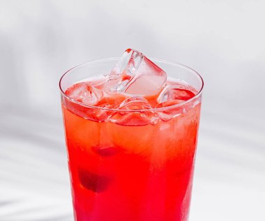 Beyaz arkaplanda palmiye yaprağı gölgesi olan beyaz küpün üzerinde duran buzlu kırmızı kokteyl.