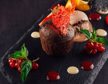 Çikolatalı lav keki, kırmızı frenk üzümü, nane ve çilekle süslenmiş bir top dondurmayla siyah taştan bir tabakta servis edilir.