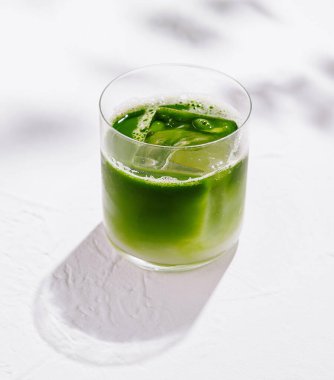 Buzlu matcha latte, arka planda yaprakların gölgeleri olan beyaz bir masada oturuyor.