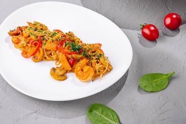 Karides ve sebzeli Udon eriştesi doğranmış yeşillikler ve gri arka planda duran beyaz tabakta susam tohumları.