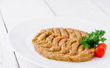 Maydanoz ve domateslerle süslenmiş beyaz bir tabakta beyaz ahşap bir masada duran Baba Ghanoush.