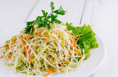 Parçalanmış beyaz lahana, rendelenmiş havuç ve beyaz tabakta dereotu ile taze salata, ahşap masada servis edilir.