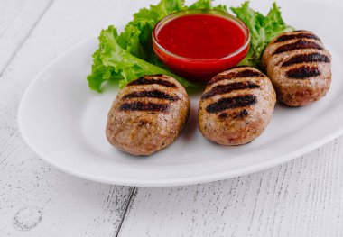 Kırmızı sos ve marullu beyaz bir tabakta üç sulu ızgara köfte servis edilir.