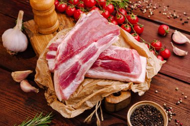 Domuz kaburgaları, olgun domatesler, sarımsak karanfilleri ve ahşap bir biber değirmeninin yanında buruşmuş kağıda konur ve masada kırsal bir pişirme atmosferi yaratır..