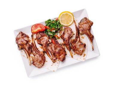 Sulu kuzu pirzolaları beyaz dikdörtgen bir tabakta, canlı otlar, bir dilim limon ve leziz bir yemek sergilenen ızgara domateslerle birlikte düzenlenir..