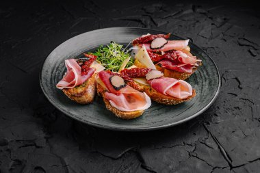 Sanatsal olarak hazırlanmış şarküteri tabakları, peynir dilimleri ve yeşilliklerle birlikte prosciutto ve kurutulmuş domatesler de dahil olmak üzere çeşitli pişirilmiş etleri sergiliyor. Tabak koyu dokulu bir tabakta yatıyor..