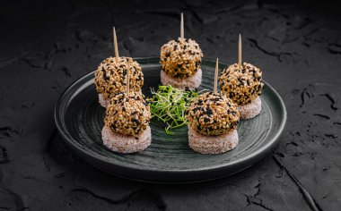 Altı susamlı top, küçük ekmek kabukları üzerinde duran siyah bir tabakta sıralanır. Taze yeşillikler sanatsal bir şekilde sunuluyor ve toplantının genel cazibesini artırıyor..