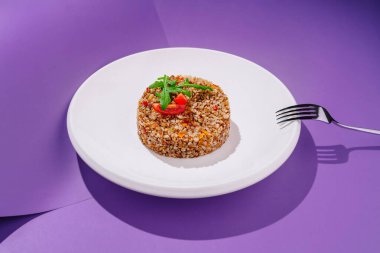 Güzel düzenlenmiş bulgur salatası beyaz bir tabakta, domates dilimleri ve taze otlarla süslenmiş. Mor arkaplan rengarenk bir kontrastlık ekler ve yemeğin cazibesini arttırır..