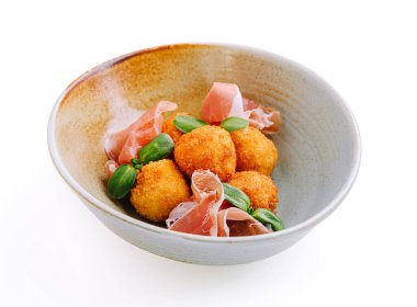 Bir kase altın kahverengi çıtır peynir toplarını sunar. Yanında da lezzetli jambon ve taze otlar bulunur. Gurme mutfak sunumunu vurgular..