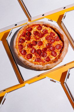 Yeni pişmiş pepperonili bir pizza, etrafına düzgünce yerleştirilmiş boş pizza kutularıyla canlı sarı bir yüzeye oturur. Sıcak renkler iştah açıcı görünümü arttırır..