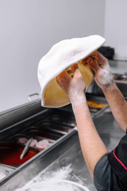 Canlı bir mutfakta aşçı uzmanlığını, taze pizza hamurunu havaya fırlatarak sergiliyor. Mutfak faaliyet ve leziz kokularla uğuldarken un ellerini tozlar..