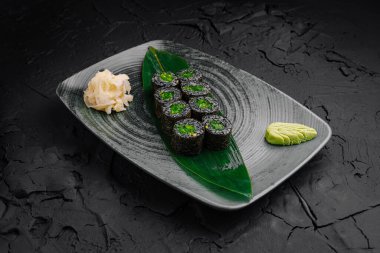 Wasabi ve zencefil turşusuyla dokulu gri bir tabakta sergilenmiş canlı bir vejetaryen suşi rulosu. Yemek geleneksel Japon aşçılık sanatını gözler önüne seriyor..