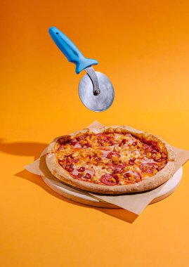 Peynirli ve pepperonili lezzetli bir pizza parlak turuncu arka planlı ahşap bir tahtanın üzerinde oturur. Mavi bir pizza kesicisi havada süzülüyor, pizzayı dilimlemek üzere..