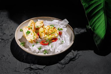 Bir tabakta, dilimlenmiş domatesler ve taze ekmekle çevrili kremalı bir peynir sergileniyor. Yemek zarif bir şekilde siyah bir yüzey üzerindeki bitkilerle düzenlenmiş..