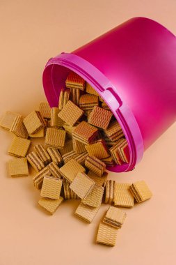 A pink container rests on its side with numerous golden waffle biscuits scattered across a peach-colored background. This display creates a playful and inviting setting. clipart