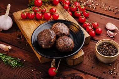 Üç mükemmel pişmiş köfte, vişneli domatesler ve aromatik otlarla çevrili siyah bir tabakta rahat bir yemek için ideal ahşap bir yüzeye kurulmuş..