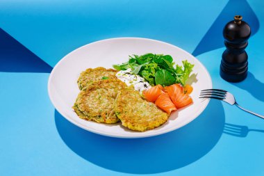 A plate of zucchini fritters is presented alongside smoked salmon and a fresh salad of leafy greens. The vibrant blue backdrop adds a lively touch to the meal, creating an inviting atmosphere. clipart