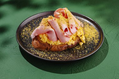 Lezzetli kahvaltı, üzerinde jambon dilimleri ve kızarmış ekmekle çırpılmış yumurta var. Tabak, canlı yeşil bir zemine karşı siyah bir tabakta servis edilir..