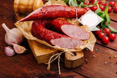 Cured sausage is displayed on a wooden cutting board alongside fresh tomatoes and garlic, showcasing vibrant colors and rich textures, creating an enticing culinary atmosphere. clipart