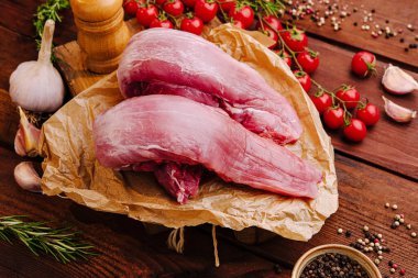 İki pişmemiş domuz filetosu sarımsak, kiraz domatesleri ve ahşap bir yüzeyde çeşitli baharatlarla çevrili kahverengi parşömen kağıdının üzerinde dinleniyor..