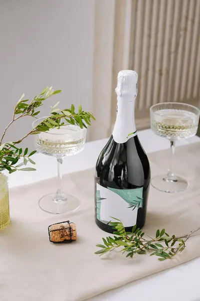Masa, bir şişe köpüklü şarap, iki zarif bardak, bir mantar ve dekoratif olarak düzenlenmiş taze yeşillikle bir kutlama için hazırlandı. Yumuşak ışıklandırma şenlikli atmosferi güçlendirir.