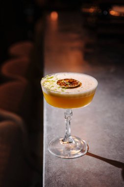 Köpüklü narenciye soslu güzel bir kokteyl ve bir dilim kurutulmuş meyve cilalı bir tezgahta duruyor. Ortam aydınlatması ayarlara sıcaklık katıyor.