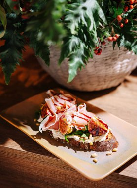 Açık yüzlü bir sandviçte taze yeşillikler, dilimlenmiş jambon ve incirler bir tabakta güzelce sunulur. Dekoratif bir bitki kırsal ahşap masa düzenine cazibe katar..