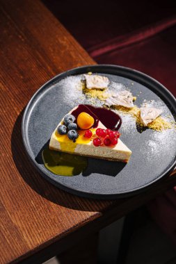 Taze böğürtlen ve renkli soslarla kaplı bir dilim kremalı cheesecake koyu bir tabakta güzelce sunuluyor. Sıcak atmosfer bu zevkli ikramların tadını artırıyor..