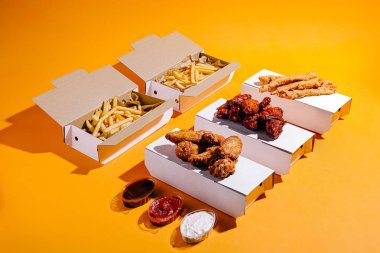 Kızarmış tavuk parçaları ve altın kızartmalar, parlak turuncu bir yüzey üzerinde, kutuların yanına soslar yerleştirilmiş, paket servis kutularında düzenlenir..