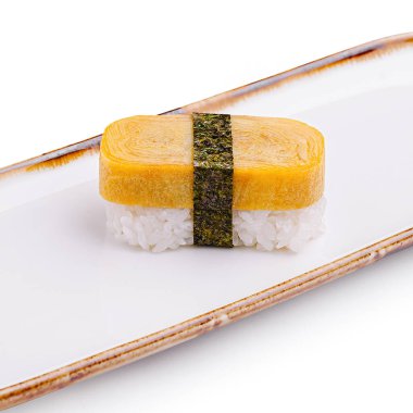 Çok güzel işlenmiş bir suşi parçası, yapışkan pirincin etrafına sarılmış, kırsal beyaz bir tabakta zarif bir şekilde sergilenmiş, mutfak sanatını gözler önüne seren tamago omletinden oluşur..