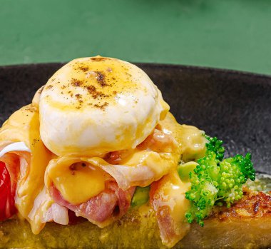 Ağız sulandıran bir kahvaltı tabağında jambon, erimiş peynir ve taze sebzelerden oluşan bir yatakta haşlanmış yumurta sergileniyor. Canlı renkler ve zengin dokular bu tatmin edici yemeği vurgular..