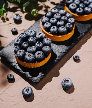 Üç canlı yabanmersinli tart taze yaban mersiniyle çevrili siyah bir levhada otururlar. Zarif düzende tatlıların zengin renkleri ve dokuları vurgulanıyor..