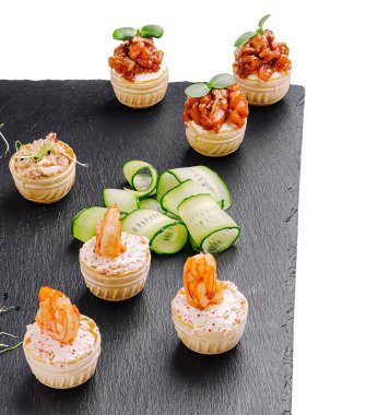 Bir dizi lezzetli deniz ürünleri aperatifleri karanlık bir tabakta cazip bir şekilde duruyor. Karides ve tavuk küçük hamur bardaklarında servis edilir, yeşilliklerle süslenir ve yanında salatalık dilimleri ile birlikte..