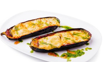 İki fırında patlıcan dilimi erimiş peynir ve canlı yeşil bitki sosuyla kaplıdır. Bu lezzetli yemek, iştah açıcı görünümünü gösteren beyaz bir tabakta sunuluyor..