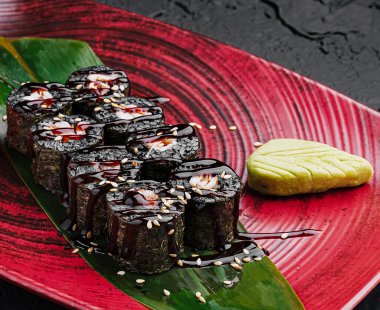 Suşi ruloları kırmızı oval bir tabağa güzelce yerleştirilmiş, üzerine sos serpiştirilmiş ve susam tohumları serpilmiş. Bir porsiyon wasabi yeşil yapraklı ruloların yanına konur..