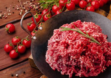 Cömert bir porsiyon taze kıyma, parlak kiraz domatesleri ve biberiye filizleriyle çevrili koyu bir tavaya konur. Kırsal ahşap masa davetkar atmosferi güçlendirir..