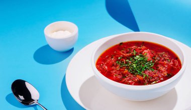 Bir kase kırmızı Ukrayna çorbası beyaz bir tabağa yerleştirilmiş taze otlarla süslenir. Küçük bir tabak krema ve gümüş bir kaşık parlak mavi bir yüzey üzerinde dinleniyor..