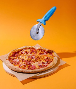 Lezzetli bir pepperonili pizza, parlak turuncu arka planlı ahşap bir kesme tahtasının üzerinde oturur. Bir pizza kesicisi havada süzülüyor, lezzetli sunumu dilimlemeye hazır..
