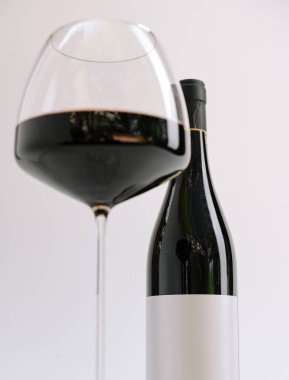 Güzel şekilli bir kırmızı şarap kadehi, zarif bir şarap şişesinin yanına yerleştirilmiş, zengin, koyu şarapla doludur. Arka plan basit ve dağınık, şarabın cazibesini vurguluyor..