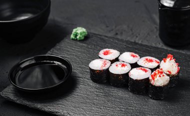 Üzerinde kırmızı susam tohumları olan renkli suşi ruloları desenli siyah bir tabakta güzelce düzenlenir. Soya soslu küçük bir tabak ve bir tutam wasabi yemeği tamamlar..