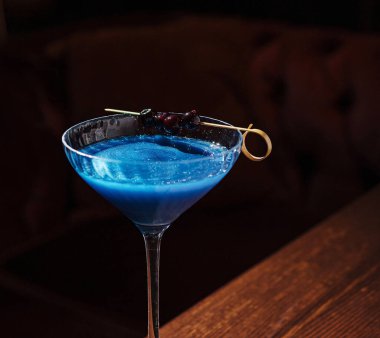 Canlı bir mavi kokteyl, böğürtlenlerle süslenmiş saplı bir bardakta zarif bir şekilde durur. İçki ışığı yansıtır, zengin rengini gösterir, koyu, rahat bir arka plana karşı..