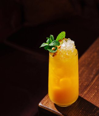 Ezilmiş buzla dolu canlı bir kokteyl ahşap bir masada duruyor. İçecek parlak turuncu, üzerinde taze nane yaprakları ve bir tutam çam fıstığı var, ferahlatıcı bir görünüm sunuyor..