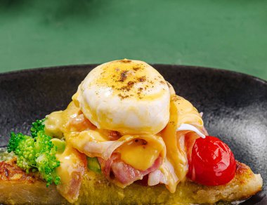 Canlı bir brunch tabağında, kavrulmuş yumurta, erimiş peynir, doğranmış jambon, kiraz domatesleri ve brokoli ile birlikte, yeşil bir arka planda, koyu bir tabakta servis edilir..