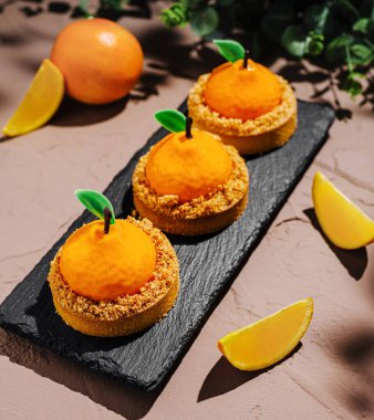 Üç portakal şekilli tatlı bir tabağa konur. Her tedavi, üzerinde kırıntılar olan parlak turuncu bir üst içeriyor. Limon dilimleri yeni bir dokunuş ekler.