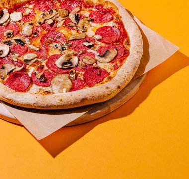 Biberli ve mantarlı yeni pişmiş bir pizza ahşap bir pizza tahtasının üzerinde canlı sarı bir zemin üzerinde duruyor ve iştah açıcı çekiciliğini gösteriyor..