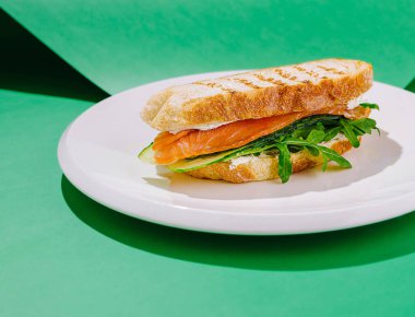 Taze yapılmış sandviç katmanları füme somon, taze roka ve ince dilimlenmiş salatalık, kızarmış ekmeğin üzerinde servis edilir. Canlı yeşil arka plan çanağın çekiciliğini arttırır.