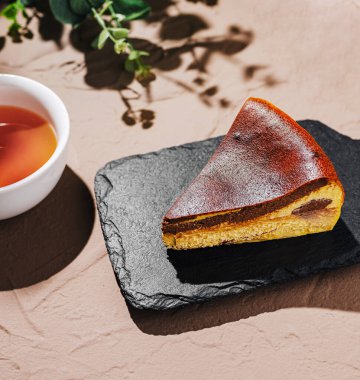 Bir dilim kremalı cheesecake, siyah bir tabakta, buharda pişmiş çayla dolu küçük beyaz bir kasenin yanında dinlenmek için mükemmel bir tatlı hazırlıyor..