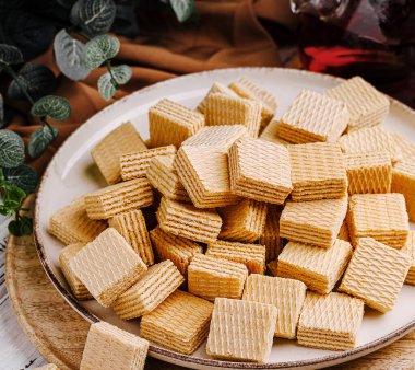 Bir tabak rahat bir ortamda düzgünce istiflenmiş gofretlerle doludur. Gofretler altın kahverengidir ve yumuşak kumaş ve yeşil aksanlarla çevrili desenli bir yüzeye sahiptir..