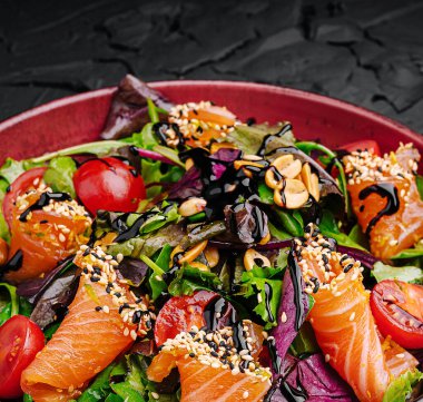 Renkli bir salatada taze somon parçaları, karışık yeşiller, kiraz domatesleri ve siyah desenli bir yüzeye aromalı sos serpiştirilmiş susam tohumları yer alır..